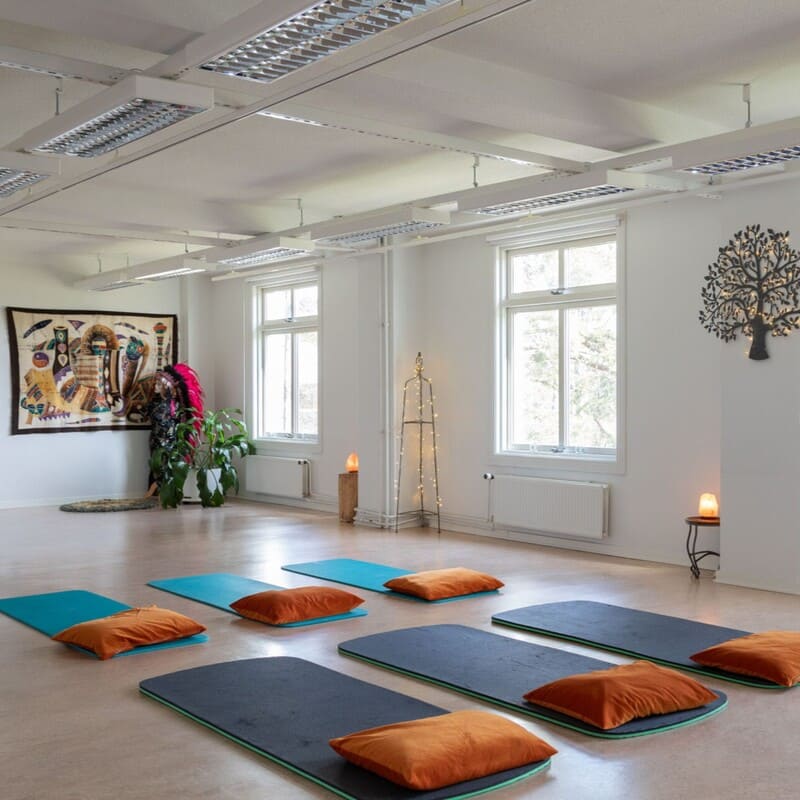 Joogamattoja ja meditaatiotyynyjä valoisassa salissa suurten ikkunoiden edessä